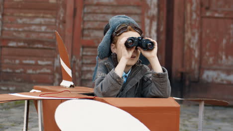 Happy-Little-Boy-With-Red-Hair-In-Hat-And-Glasses-Sitting-Outdoors-In-Wooden-Toy-Model-Of-Airplane-And-Looking-In-Binoculars