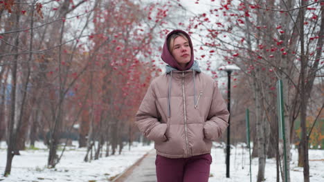 young woman wearing warm hooded jacket walks along snowy path with hands in pockets, looking thoughtful, red-berried trees line peaceful winter park