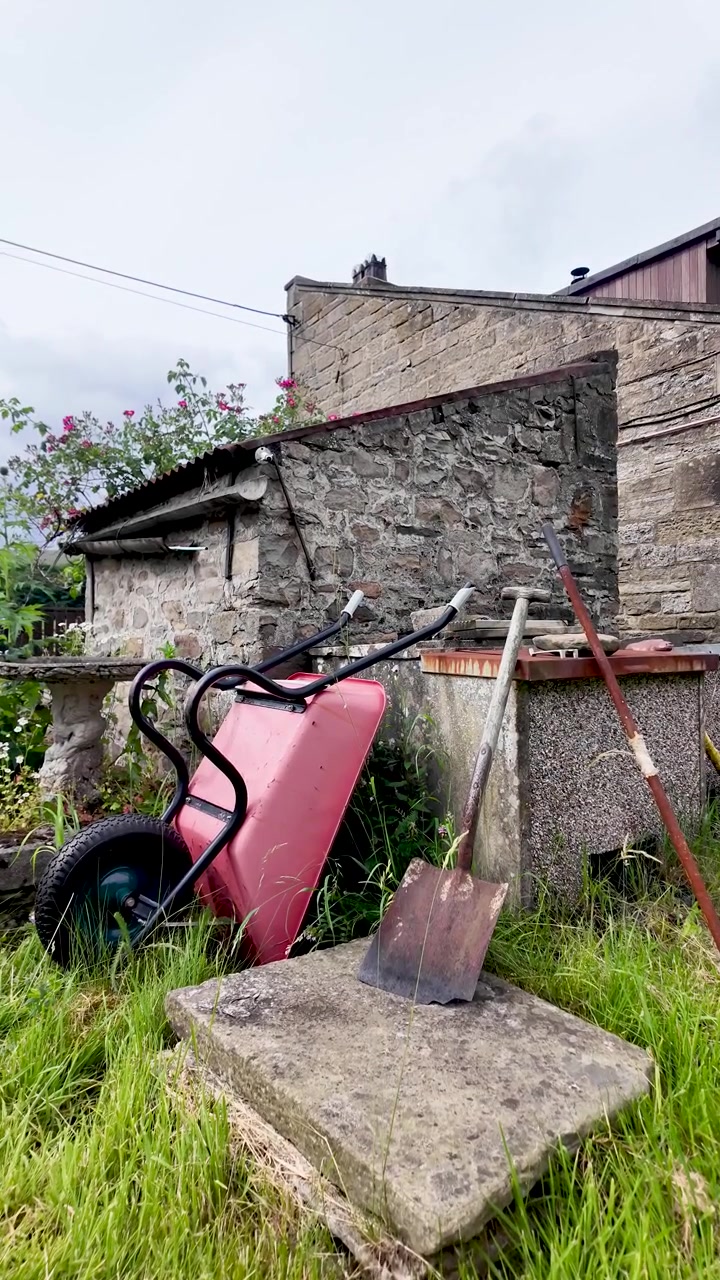 Carriola arrugginita e pale giacciono nell'erba alta accanto a una vecchia casa in pietra in un tranquillo cortile rurale. Video verticale.