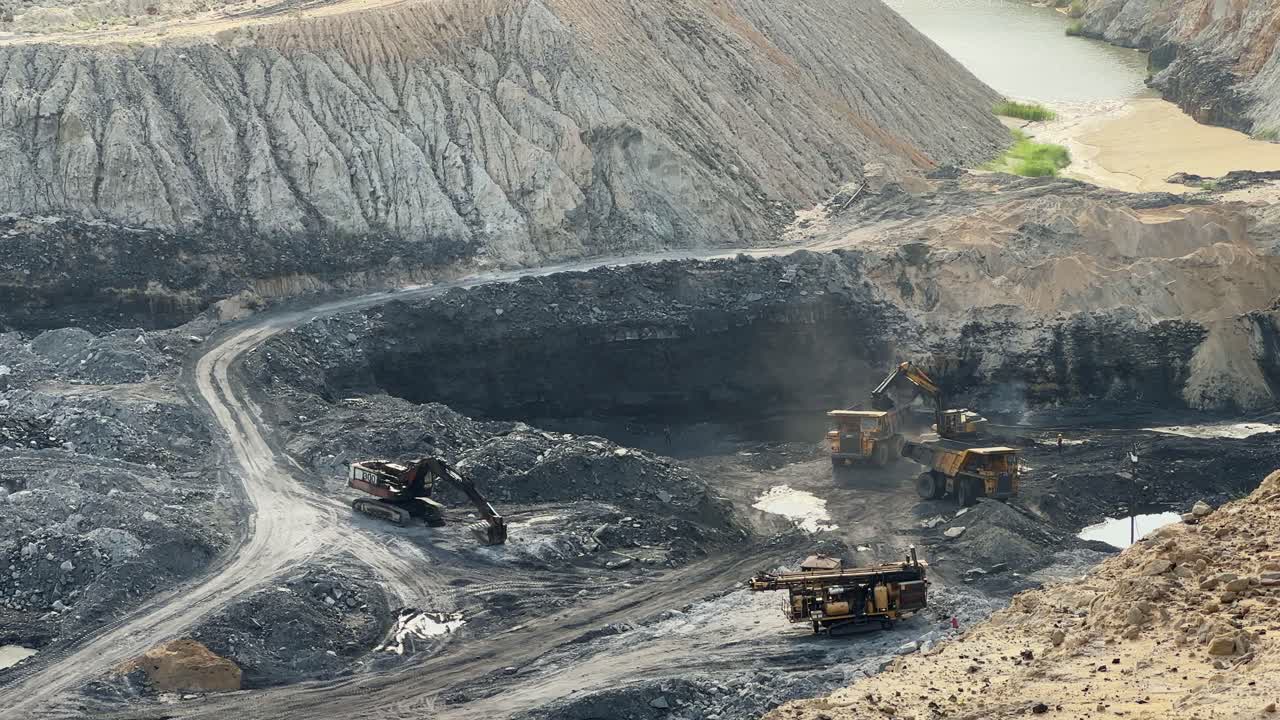 A long shot of a mining operation, a backhoe is shown loading coal into a large dump truck. Dump trucks are essential for transporting raw materials like coal in quarries and mines