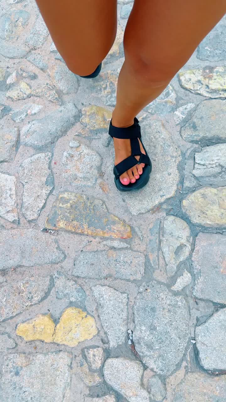 Antik Arnavut kaldırımlı sokakta siyah sandaletlerle yürüyen kadın. Engebeli gri taş kaldırımda siyah spor sandaletli ayakların yukarıdan görünümü.