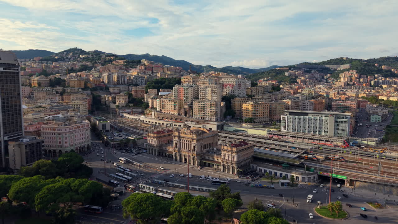 Drone advances toward the railway tracks and station in Genoa at sunset, showing traffic, trees, a tall modern building, surrounding city, and mountains in the background