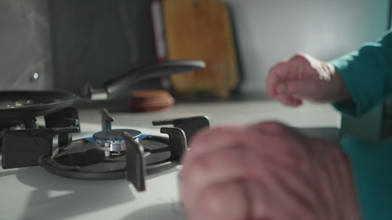 Elderly woman in green sweatshirt cooking on gas stove, flipping pancake with pan. Modern kitchen with sleek surfaces and gentle lighting, capturing independence in daily cooking routine
