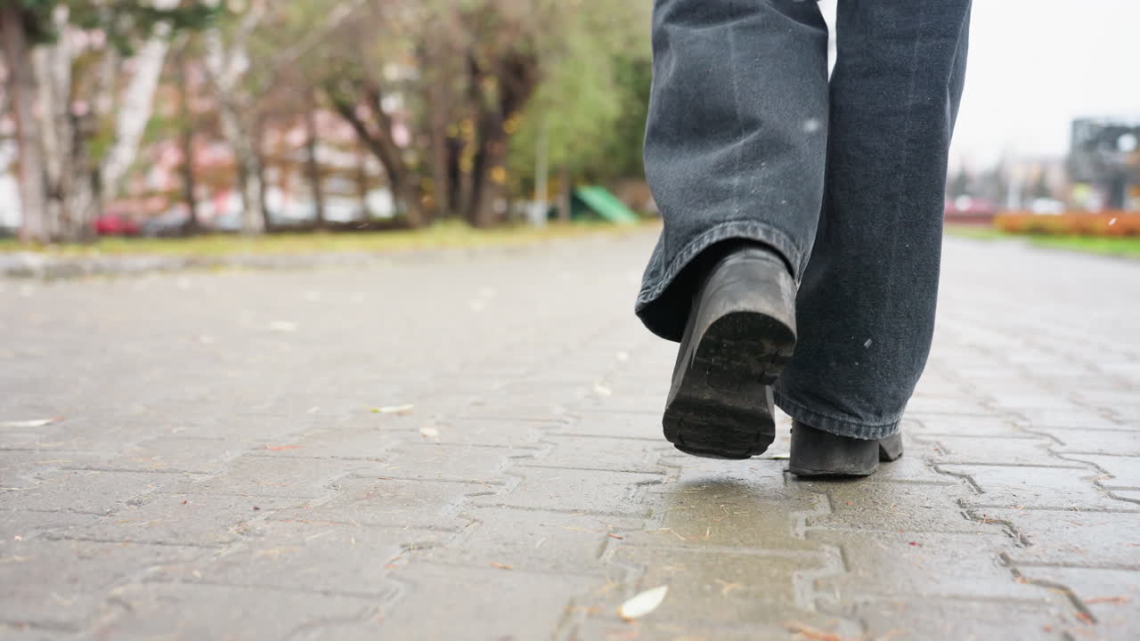 A human leg wearing black trousers and black boots walking slowly on outdoor paved path during calm overcast day, capturing foot movement and subtle step motion in quiet urban environment