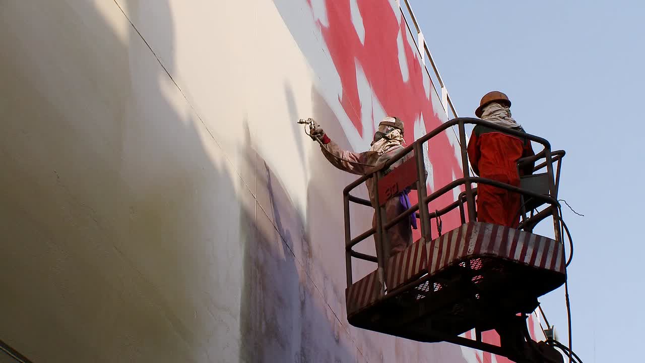 Workers in a cherry-picker spray-painting the side of an oil tanker in dry dock