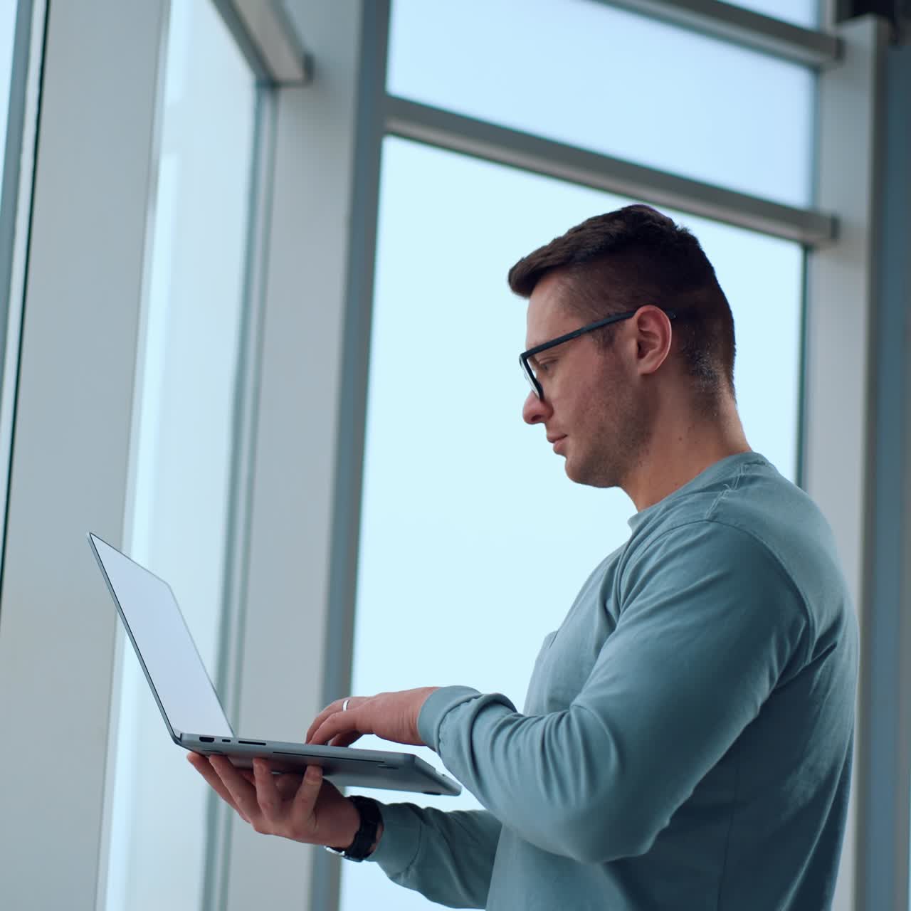 Serious confident businessman standing indoors neat the big window. Man opens his laptop typing something on the keyboard. Side view