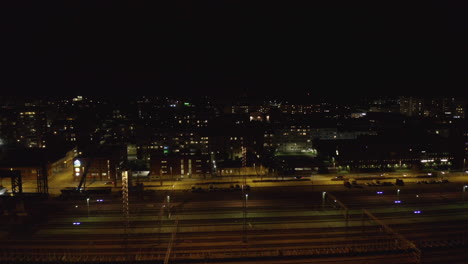 Drone-shot-of-empty-railways-depot-and-cars-drive-on-city-street-at-night