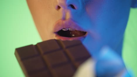 disparo de cerca de los labios de una hermosa mujer joven disfrutando y lascivamente tomando un bocado de un delicioso chocolate contra un fondo verde con contraste azul en su cara en cámara lenta