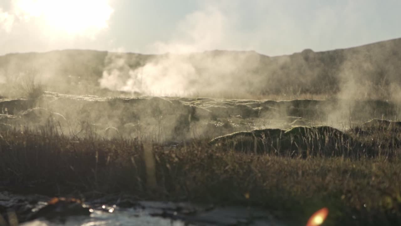 An atmospheric wide shot captures the geothermal area of Geysir in Iceland, with significant amounts of steam rising from various hot springs and vents across the landscape.