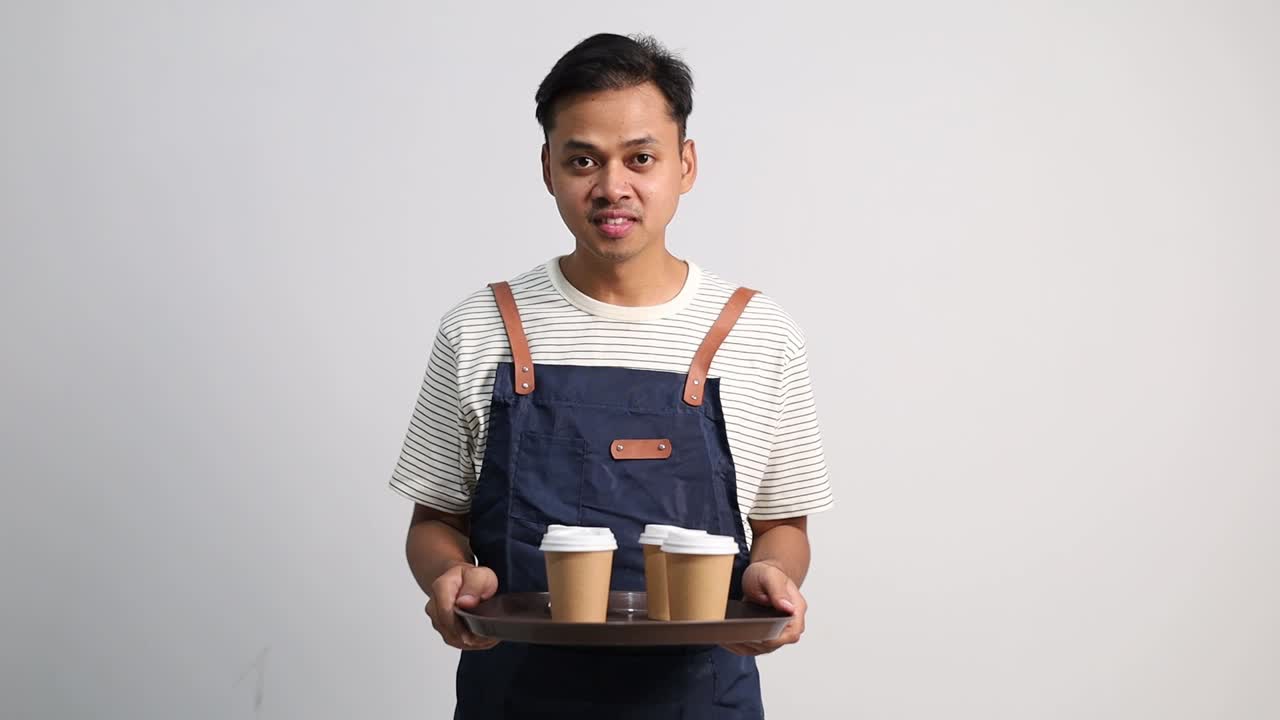 Seorang pelayan kedai kopi pria mengenakan celemek sambil membawa nampan berisi gelas kopi kertas dengan gerakan menyerahkan, mengantarkan, atau memberikan pesanan minuman kepada pelanggan
