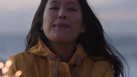 섣달 그믐날을 축하하는 불꽃놀이를 들고 있는 행복한 아시아 여성의 초상화를 닫고 일몰 때 해변에서 독립 기념일 축하를 즐기며 웃고 있습니다