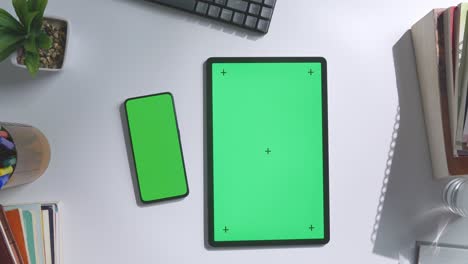 top down view of a laptop computer with mock up green screen chromakey display on a wooden office desk next to notebook with pens, glasses, and a glass of water. slow zoom out, close up