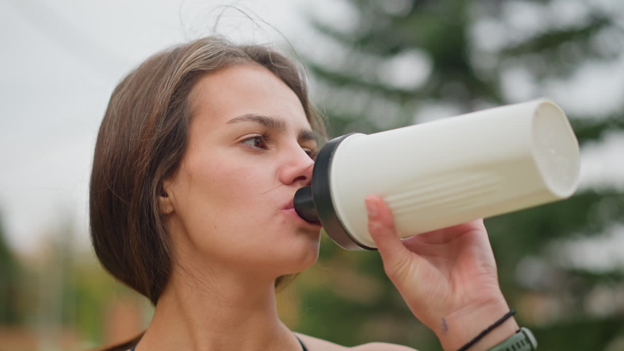 Close-up view of an athlete drinking water from black and white water bottle, focusing on hydration and recovery during outdoor workout session, emphasizing fitness and wellness routine