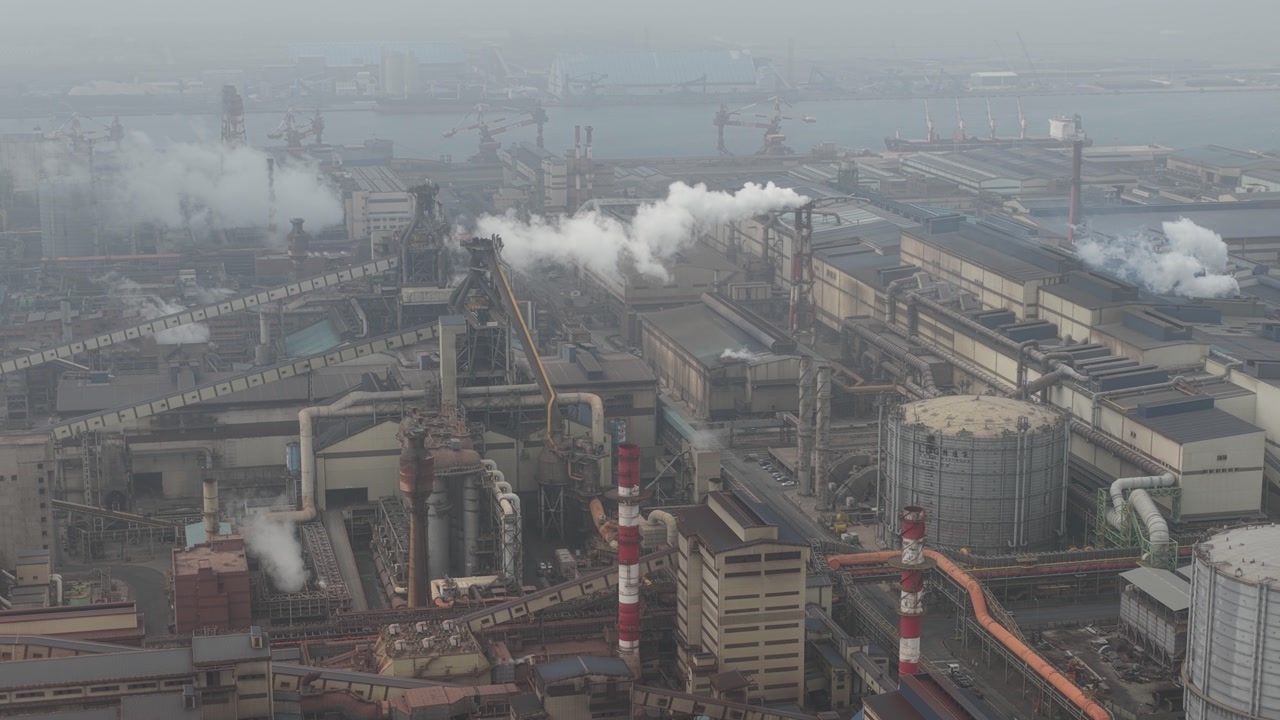 Vast industrial complex with numerous smokestacks releasing plumes of smoke and steam into the hazy sky, highlighting the scale of heavy industry and its environmental impact on a foggy day