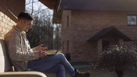 caucasian man having a video call on laptop while sitting on a bench outside a country house