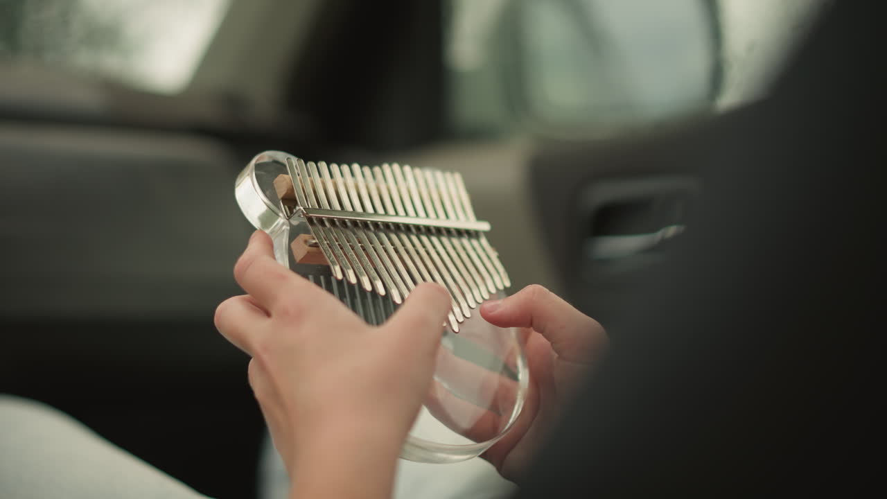 Pasajero tocando la kalimba en el coche; joven artista ensayando para tocar en la calle; luz cálida del sol entrando por la ventana; primeros planos de las manos; vaqueros informales; alegre tintineo; práctica durante el viaje; concentración máxima.
