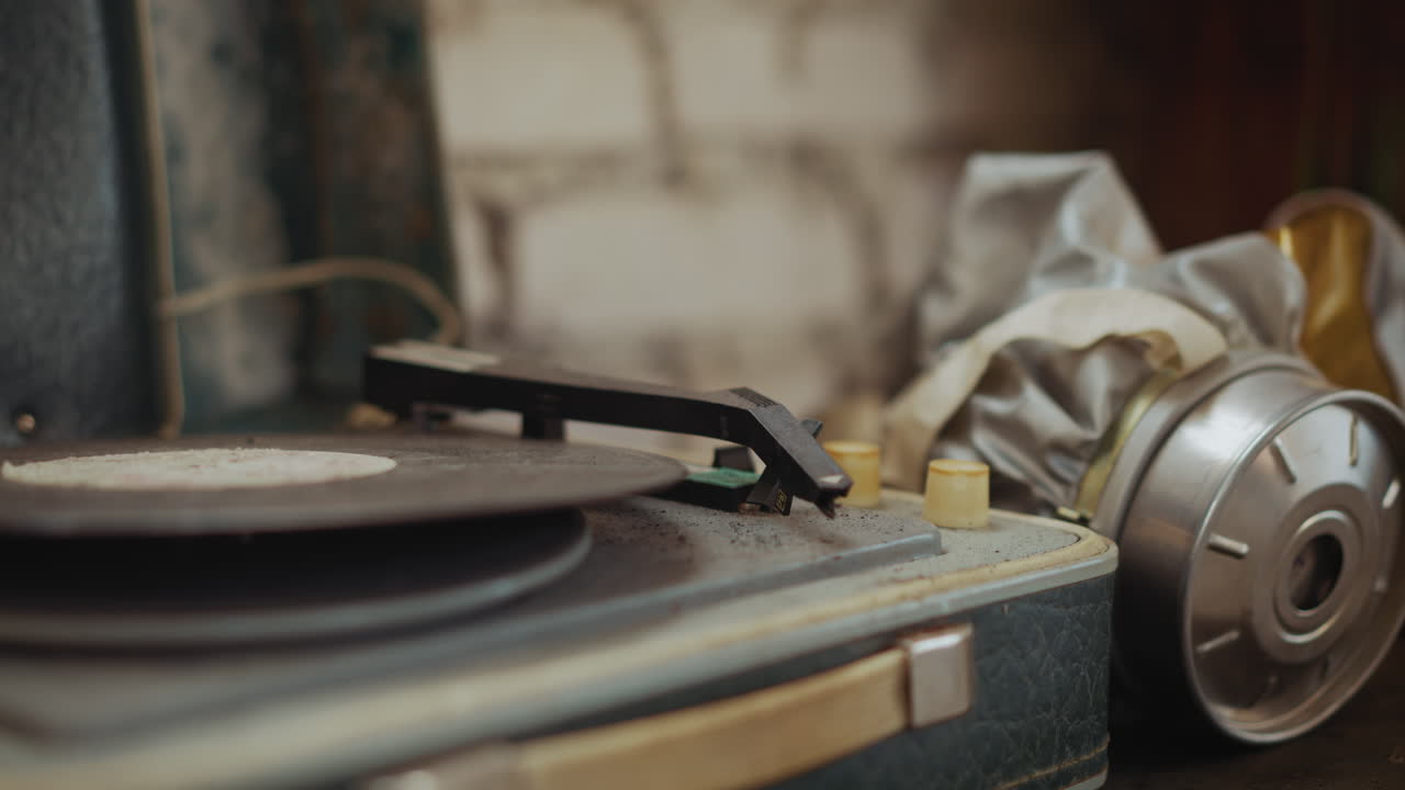 Close up of person take dusty vinyl record on vintage turntable beside silver gas mask with yellow visor, scene evokes post disaster survival
