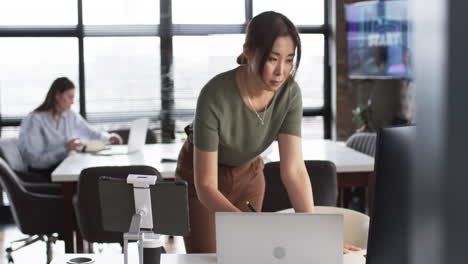 Asian-elder-works-on-laptop,-young-Caucasian-peer-in-back