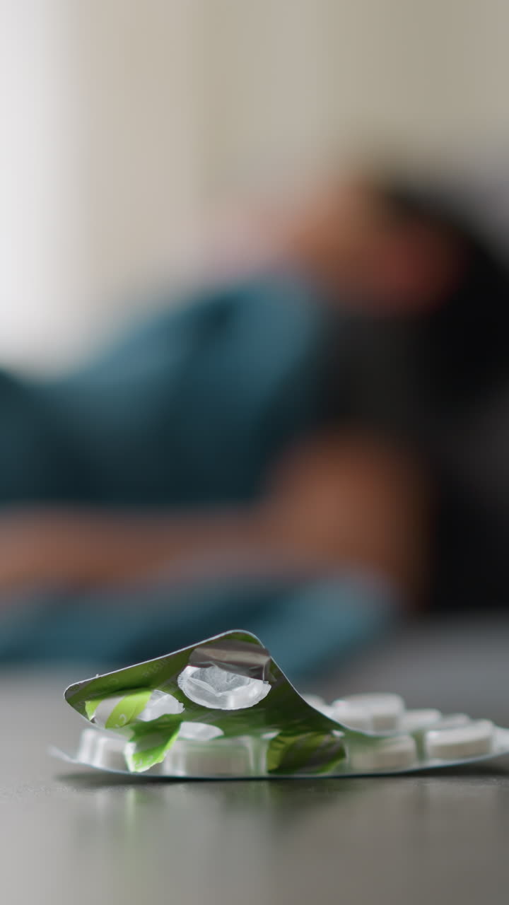 Close-up of medicine and cup on table with blurred view of someone adjusting on bed, relaxing, peaceful setting, possibly indicating a sick day or recovery period