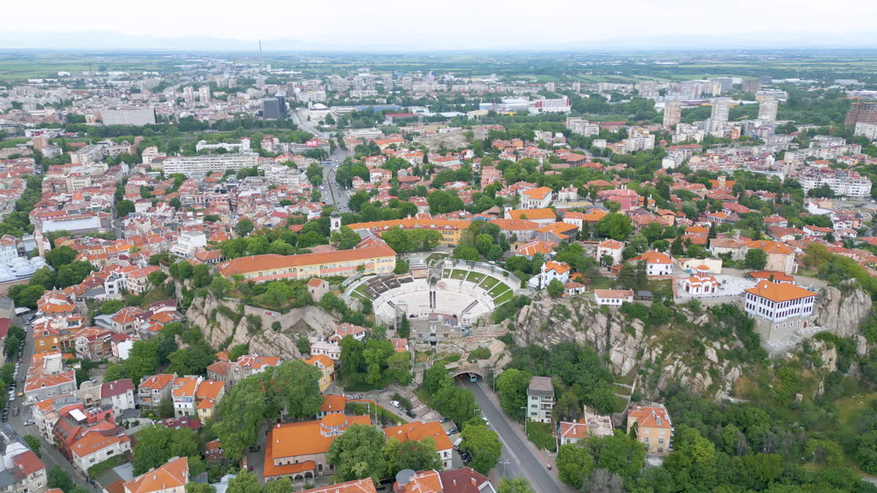 Pullback aerial reveals the Ancient Theatre of Plovdiv, widening from marble seating to the cityscape and the tunnel road beneath, filmed in early spring