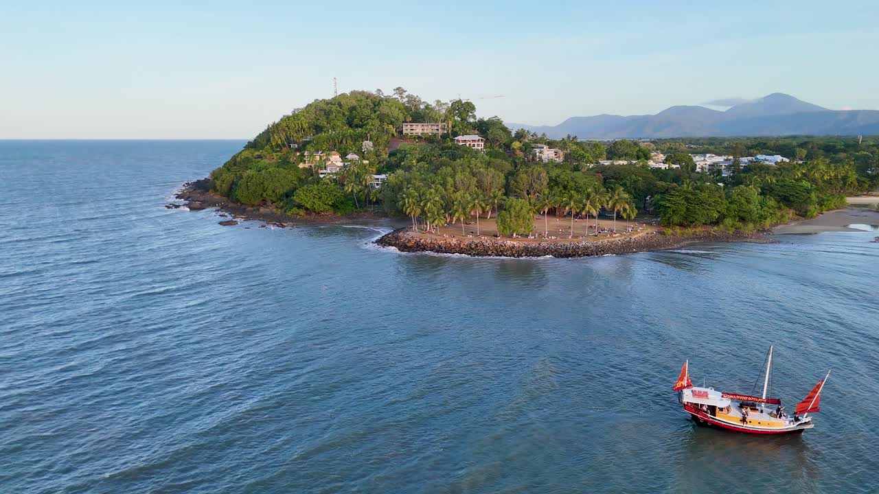 Drone captures serene coastal landscape with lush greenery and a boat approaching. Soft golden hour lighting enhances the tranquil scene