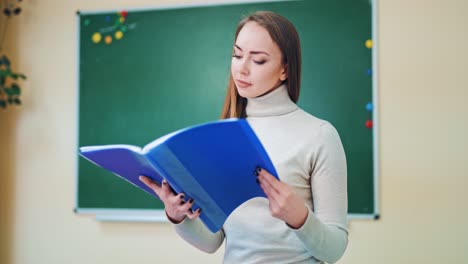 young teacher with a folder in the classroom. attractive pedagogue holds a blue folder and looks for the necessary information. educational concept.