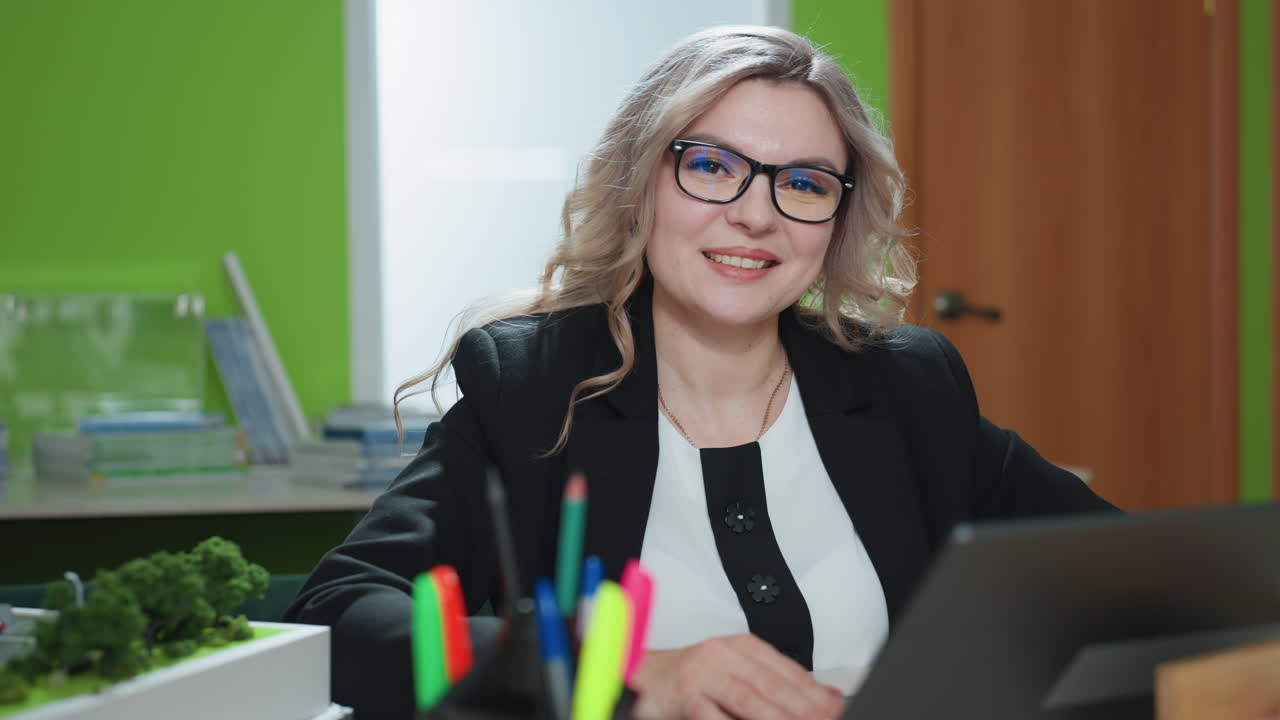 elegant lady with curly blonde hair and glasses looks up from laptop with warm smile in brightly lit office, surrounded by desk supplies, building model, indoor plant, and neat workspace atmosphere
