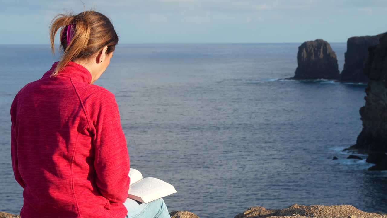 Woman reading book overlooking rocky coastal landscape during sunset