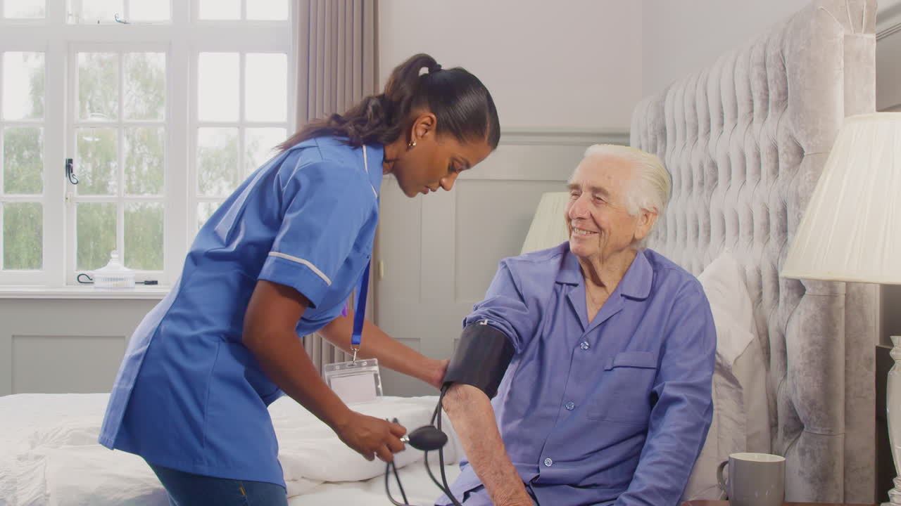 Senior Man At Home In Bed Having Blood Pressure Taken By Female Care Worker In Uniform