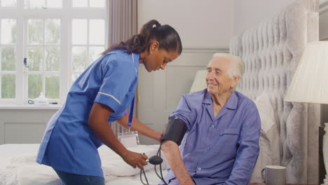 senior man at home in bed having blood pressure taken by female care worker in uniform