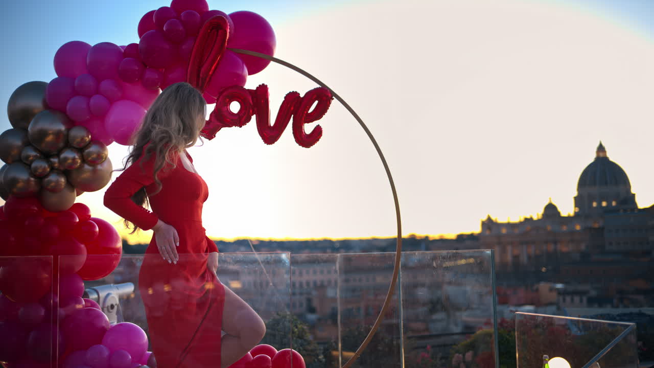 Beautiful young woman in red dress dancing at sunset near the red and pink balloon arch spelling out the word "love". Vatican city in background. Rome, Italy