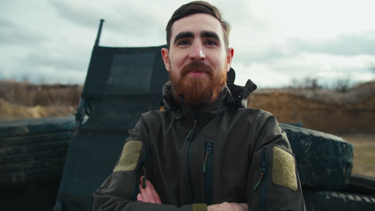 retrato de un hombre moreno confiado con barba en ropa de ejército verde oscuro cerca de neumáticos de automóviles en la estepa durante el entrenamiento de combate. hombre militar masculino confiado posando durante un ejercicio de combate fuera de la ciudad
