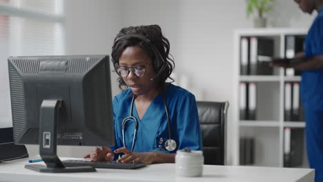 African-Female-medical-assistant-wears-white-coat-headset-video-calling-distant-patient-on-computer.-Doctor-talking-to-client-using-virtual-chat-computer-app.-Telemedicine-remote-healthcare