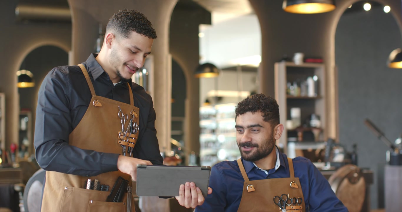 Barbieri afroamericani in grembiule che mostrano un tablet alla postazione del salone, guidando i clienti nella scelta delle opzioni di styling.