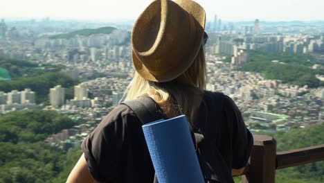 Joven-Caucásica-Con-Sombrero-Marrón-Apoyada-En-Las-Barandillas-Y-Mirando-Hacia-La-Hermosa-Ciudad-Del-Distrito-De-Seocho-gu-Desde-La-Montaña-Gwanaksan-En-Seúl,-Corea-Del-Sur