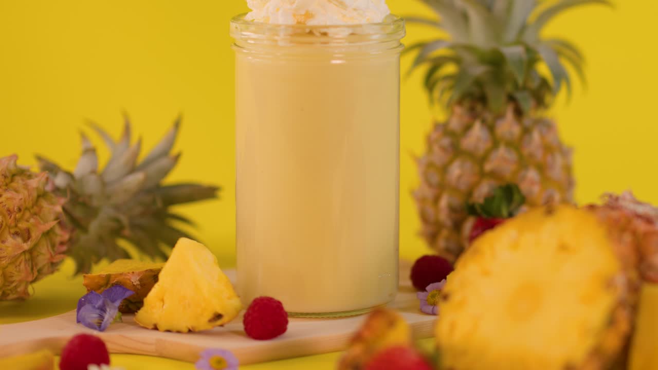 Pineapple smoothie in glass jar with whipped cream, surrounded by fruit, bright yellow background