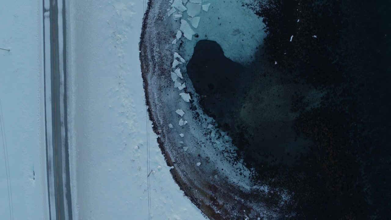 Top down aerial of snowy shoreline with small building beside dark Arctic water in winter