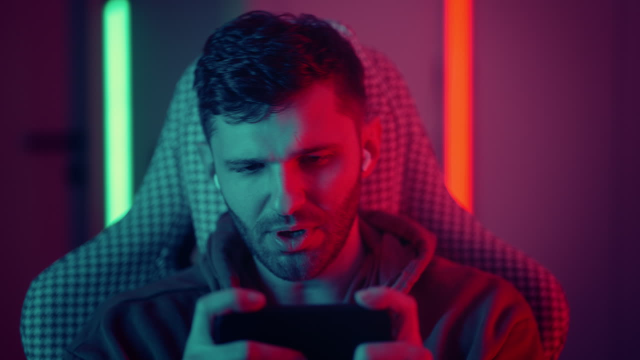Happy guy man gamer wear competition play video game online with smartphone colorful neon light in living room at night modern house. Esport streaming game online Home quarantine activity. High quality 4k footage