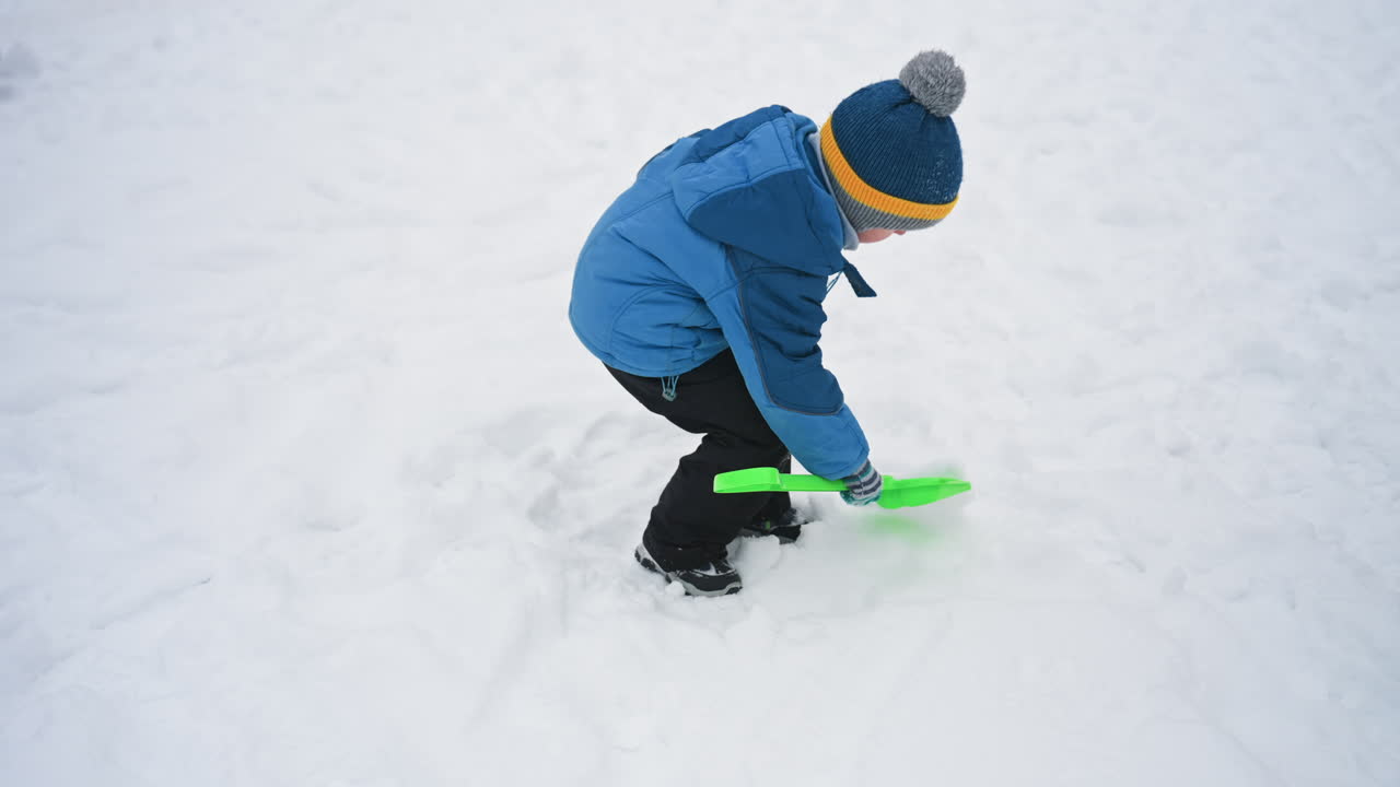 Bambino che scava la neve con una pala verde, cappotto blu e berretto di lana, concentrato a spalare neve profonda e a formare piccoli cumuli, primo piano su mani e pala con impronte sparse, scena invernale energica e autonoma.