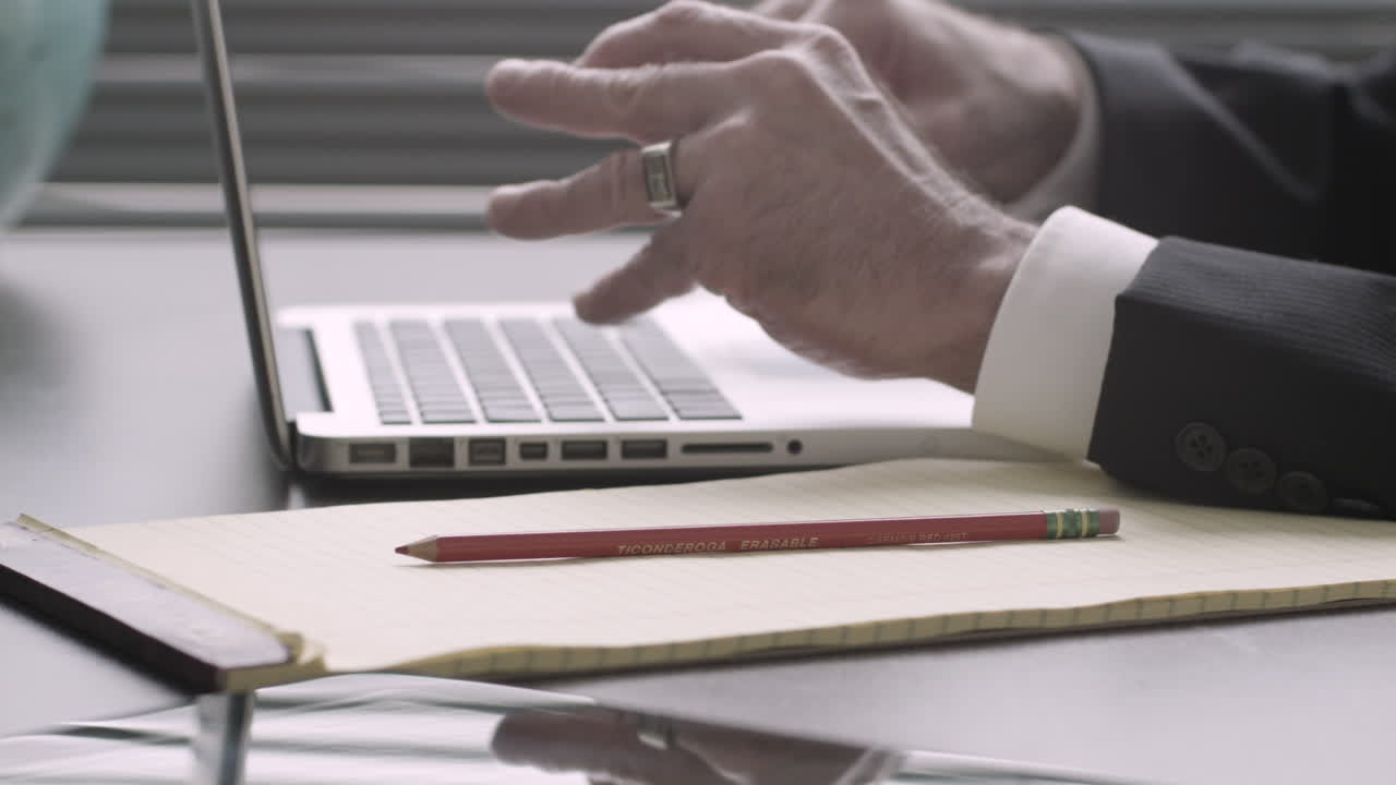 Left handed executive takes written notes and types on a laptop at his desk still shot