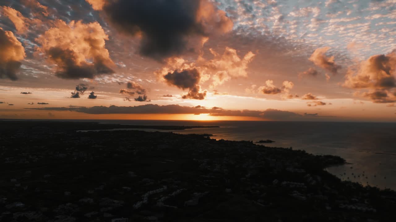 Panoramic rotating aerial footage showcasing a magnificent sunset over the dark Mauritian coastline and a scattered cityscape. The concept centers on magical island vacations, dramatic evening travel