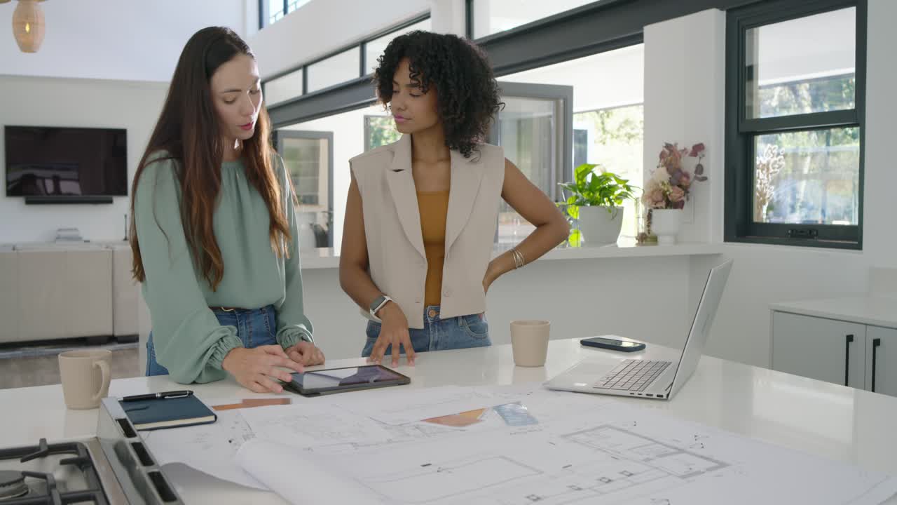 Diverse female colleagues leaning over kitchen island checking plans tapping tablet planning layout
