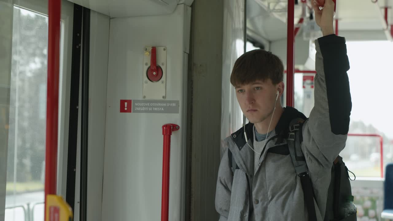 Man holds onto hand loop on trail tram bus wearing grey winter coat, listening to music