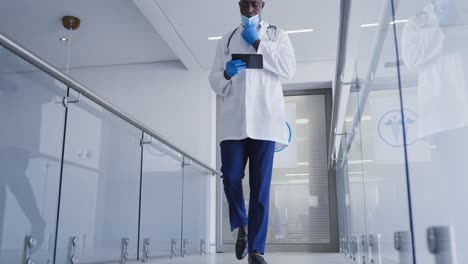 portrait of african american male doctor wearing face mask using tablet walking in hospital corridor