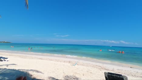 Toma-Panorámica-A-La-Izquierda-De-Una-Hermosa-Playa-Tropical-Con-Turistas-Nadando-Y-Una-Tabla-De-Windsurf-En-La-Hermosa-Playa-Del-Carmen-En-Riviera-Maya,-México-Cerca-De-Cancún-En-Un-Soleado-Día-De-Verano-De-Vacaciones