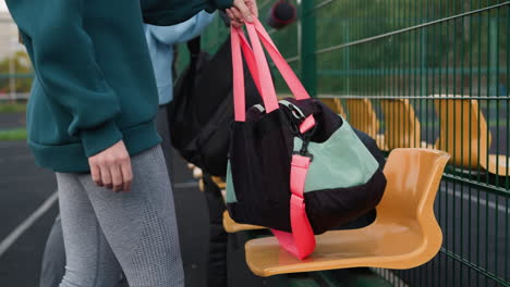 close-up of athlete walking towards sports bench, placing bag on chair near fenced outdoor court, background features yellow stadium seats, green metal fencing, and an urban sports facility