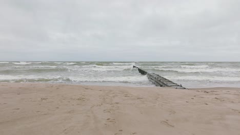 Establishing-aerial-view-of-Baltic-sea-coast-on-a-overcast-day,-old-wooden-pier,-white-sand-beach,-large-storm-waves-crushing-against-the-coast,-climate-changes,-wide-drone-shot-moving-forward-low