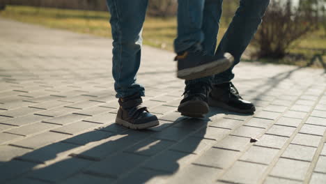 a close-up of two people s legs, one adult and one child, as they are playfully stamping there legs on the pavement together both are wearing blue jeans and sturdy boots