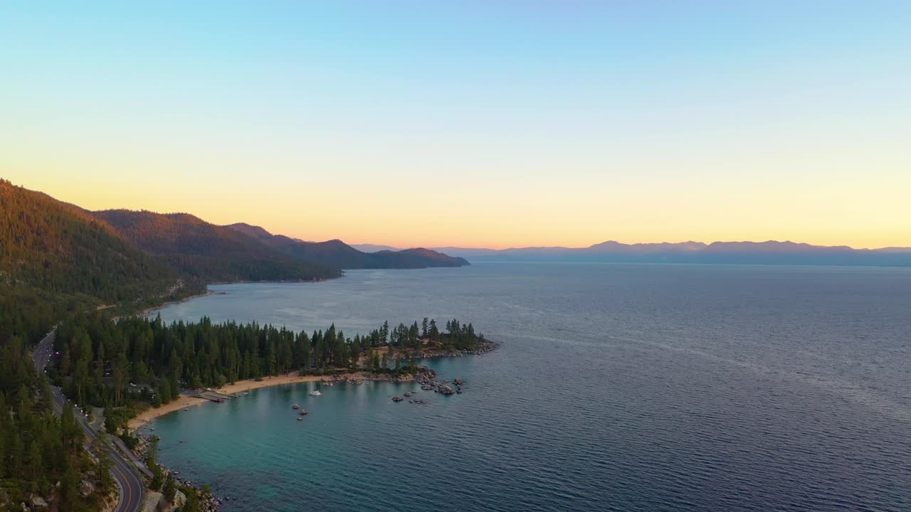 vista aérea de drones de un hermoso amanecer sobre la playa en el lago taho con una carretera o autopista que atraviesa un bosque de pinos con agua azul clara y montañas en el fondo