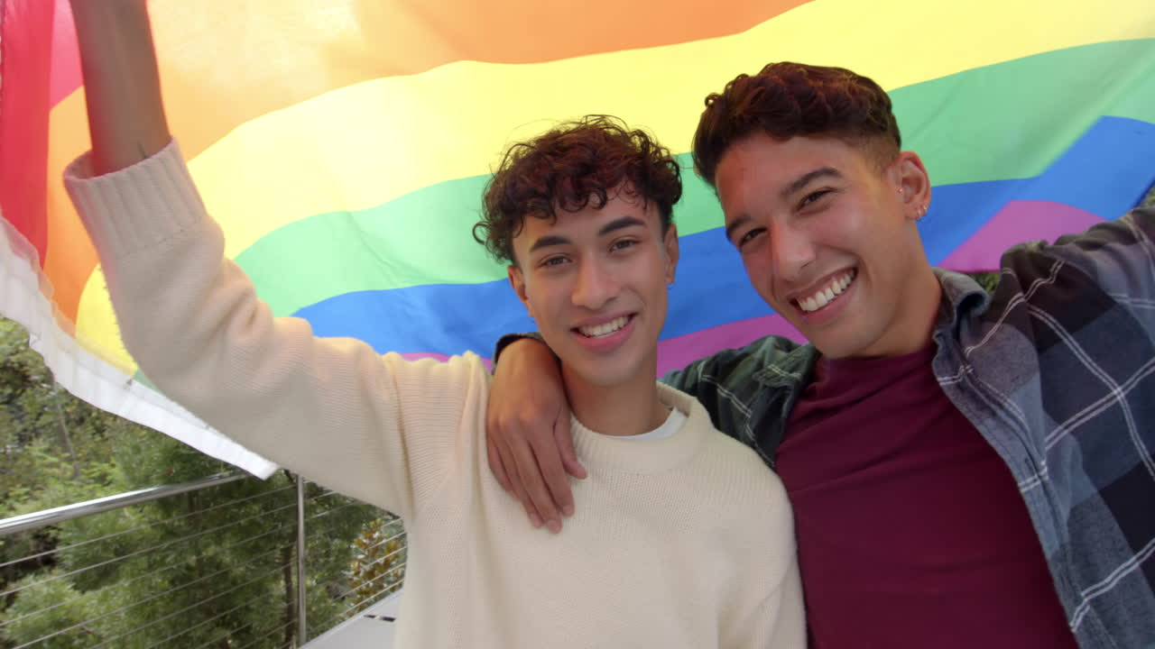 Smiling multiracial gay couple holding rainbow flag and embracing, celebrating LGBTQ+ pride, at home